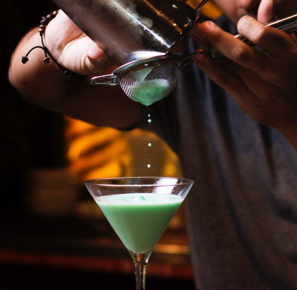 Mixologist pouring a gorgeously green tea cocktail from a shaker into a martini glass