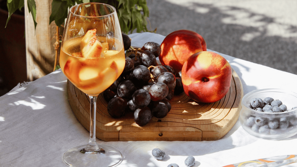 Peach iced tea in a wine glass with a chopping board next to it, covered with peaches, grapes and blueberries.